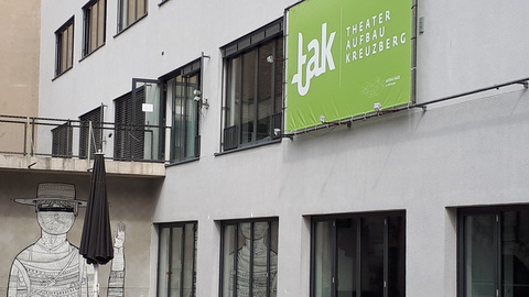 moderne weiße Betonwand mit vielen Fenstern, darauf ein hellgrünes Planenbanner mit weißen Lettern und Logo Ttak Theater Aufbau Kreuzberg