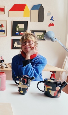 blonde, weiße Frau mit Dutt sitzt in einem blauen Pulli an einem Tisch, den Kopf auf eine Hand gestützt, sie lächelt in die Kamera, im Hintergrund farbige Bilder an der Wand