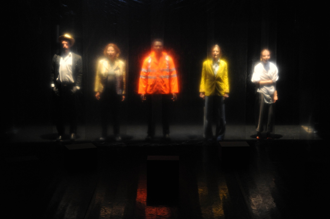 Szenenbild, eine Reihe von 5 Menschen in bunter Kleidung auf einer Bühne unscharf fotografiert