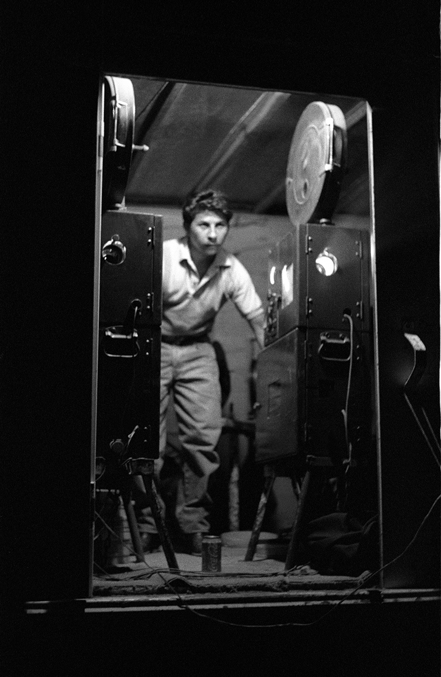 schwarz weiß Bild eines jungen Mannes in lockerer Kleidung, langärmeliges Hemd und lange Hose, zwischen zwei alten Filmprojektoren, wahrscheinlich ein Filmvorführer