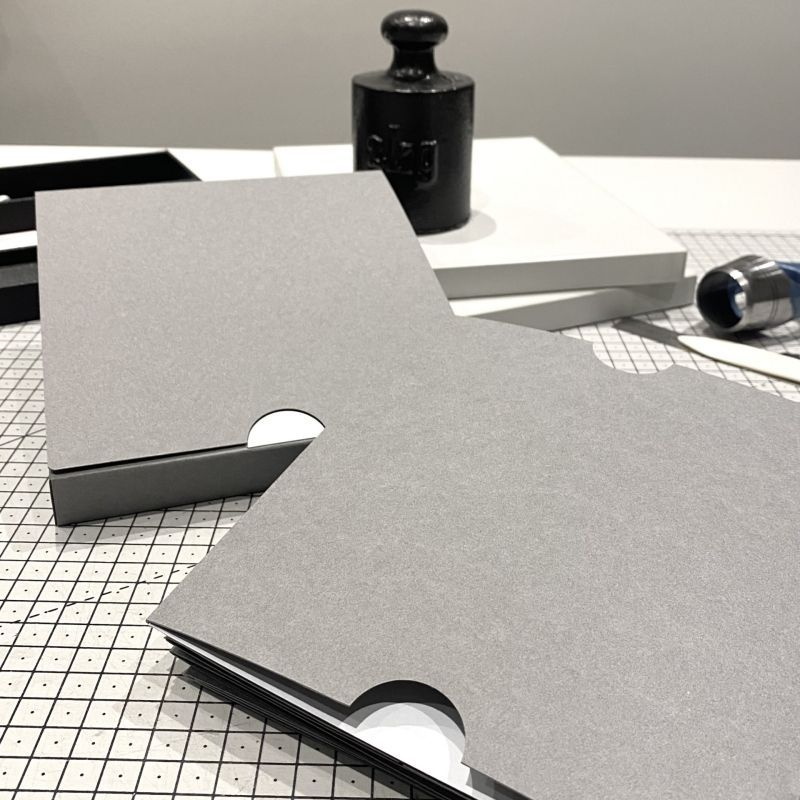 graue Kartonage mit Inhalt auf einem karierten Untergrund, im Hintergrund ein altes Gewicht zum Bücherbeschweren