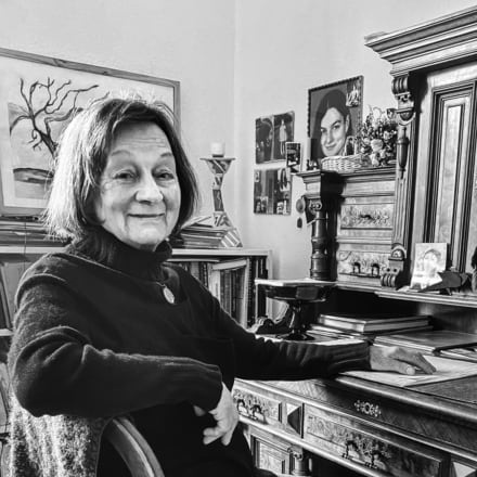 schwarz-weiß Foto einer älteren Frau sitzend an einem Sekretär, voller Raum mit Möbeln und Bildern