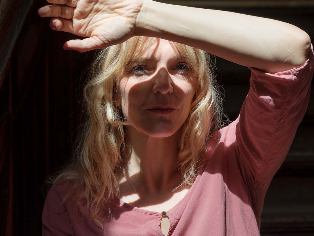 Porträt einer mittelalten weißen Frau, langhaarig blond, die in die Sonne guckt und sich den linken Arm schützend vor das Gesicht hält, sie trägt eine hellrosa-farbene, kragenlose Bluse